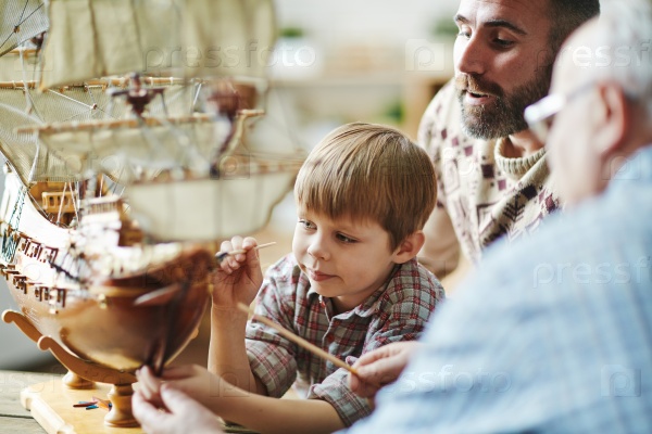 Little boy painting toy ship with his father and grandfather near by, stock photo
