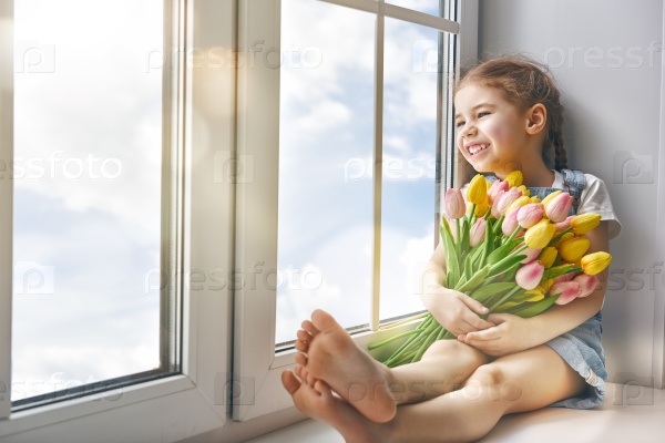 Adorable little child girl sitting on the window and holding tulips. Girl looks at the blue, pure sky and rejoices to spring and sun.