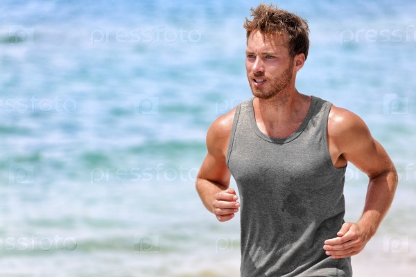 Active runner sweating running on beach. Handsome young male athlete wearing grey tank top for sweat wicking during intense cardio working on hot summer day with ocean background.