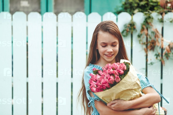 Tween girl holding a bunch of pink roses wrapped in craft paper over blue wooden fence, stock photo