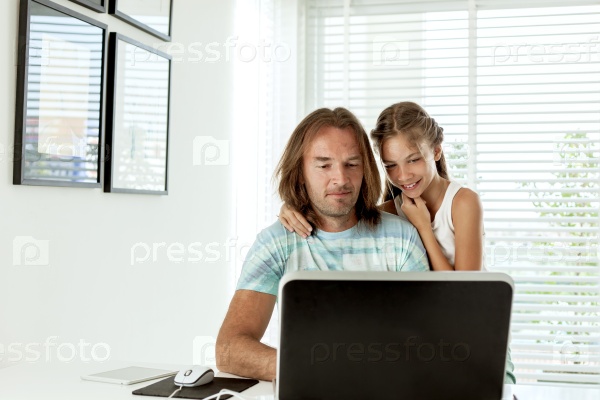 Pre teen daughter came to her dad when he was using laptop in home room. Father browsing internet with child together, stock photo