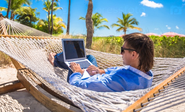 Young man working with laptop in hammock during beach vacation