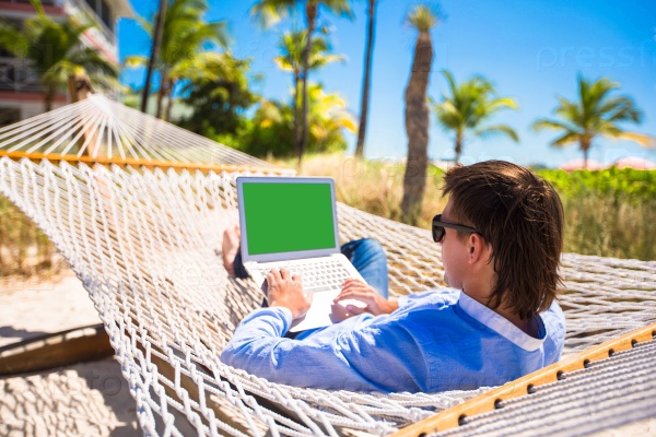 Young man working with laptop in hammock during beach vacation