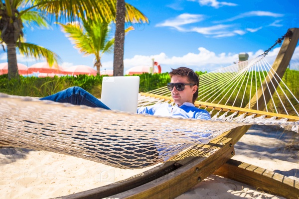Young man working on laptop in hammock at tropical beach