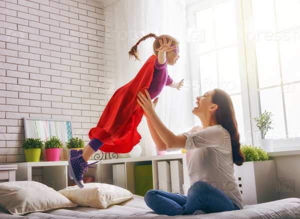 Mother and her child girl playing together. Girl in an Superman\'s costume. The child having fun and jumping on the bed.
