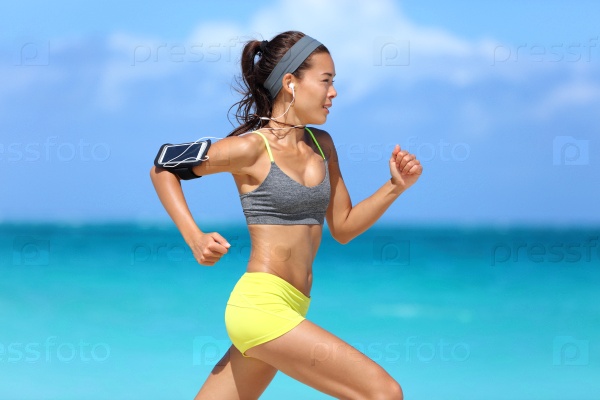 Athlete running woman runner listening to music on her phone sports armband with touchscreen and headphones earphones on summer beach. Fitness girl jogging fast training cardio and glutes.