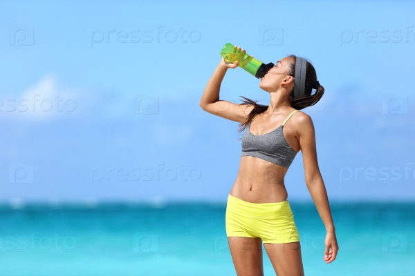 Fitness runner woman drinking water or energy drink of a sport bottle. Athlete girl taking a break during run to hydrate during hot summer exercise on beach. Healthy active lifestyle.