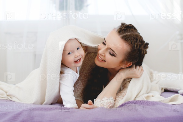 Happy family, young beautiful woman with dark hair, in a braid, gray eyes, dressed in a white lace dress, playing with his little daughter, girl - a brunette with short hair, in the bedroom on the bed with purple blanket, dampening sheets