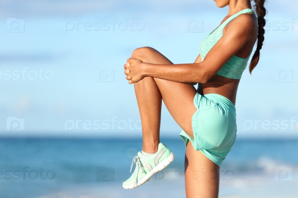 Fitness runner body closeup doing warm-up routine on beach before running, stretching leg muscles with standing single knee to chest stretch. Female athlete preparing legs for cardio workout.