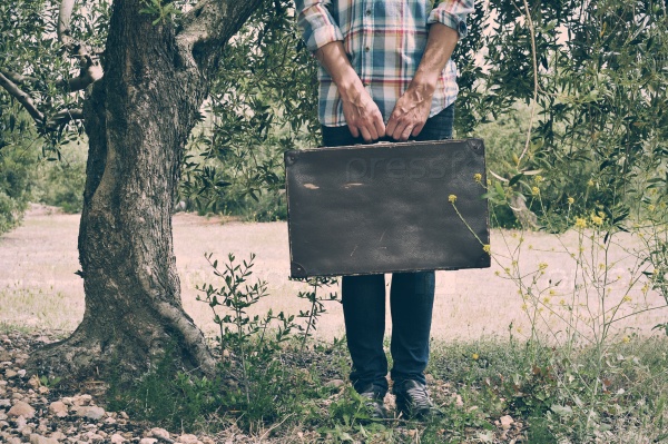 a young caucasian man wearing a plaid shirt carries an old brown suitcase in a rustic natural scenery, with a filter effect