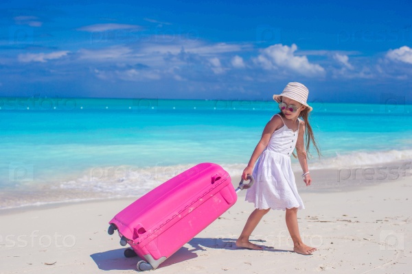 Little adorable girl with big colorful suitcase in hands walking on tropical beach