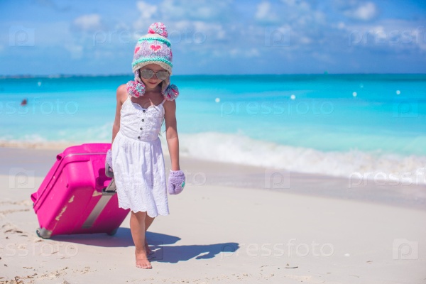 Adorable girl in warm winter hat and mittens walking with luggage on beach