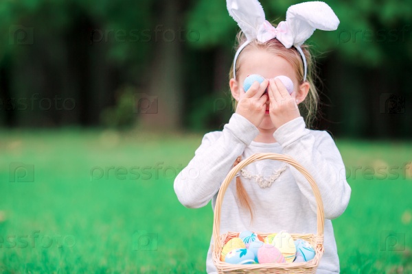 Funny little girl wearing bunny ears playing with Easter eggs on spring day outdoors, stock photo