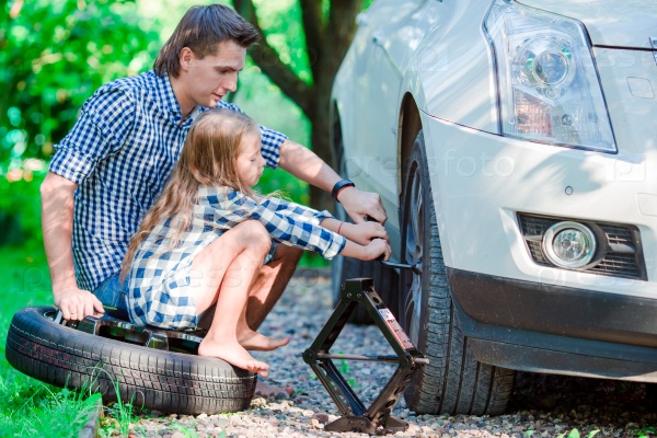 Adorable little girl sitting on a tire and helping father to change a car wheel outdoors on beautiful summer day, stock photo