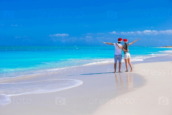 Back view of young romantic couple in red Santa hats sitting on tropical white sand beach, stock photo