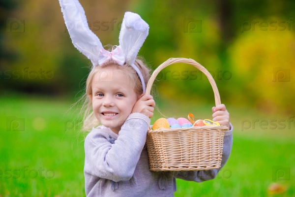 Happy little girl wearing bunny ears with a basket full of Easter eggs on spring day outdoors, stock photo