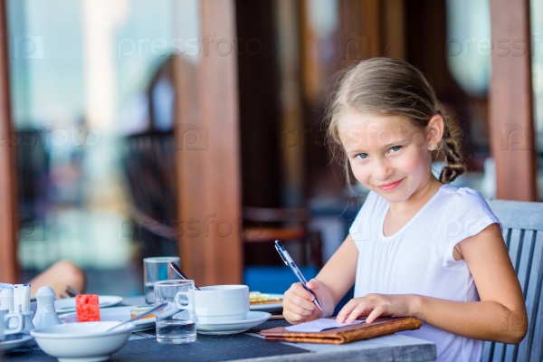 Adorable little girl signs bill after having breakfast at outdoor cafe, stock photo