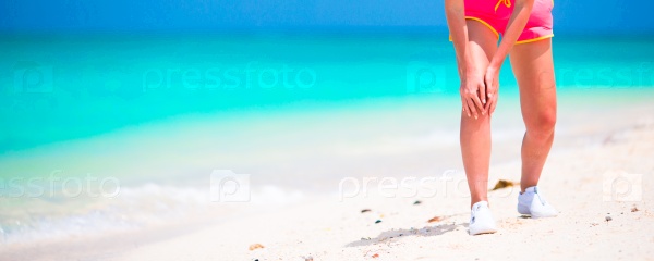 Female athlete suffering from pain in leg while exercising on white beach, stock photo