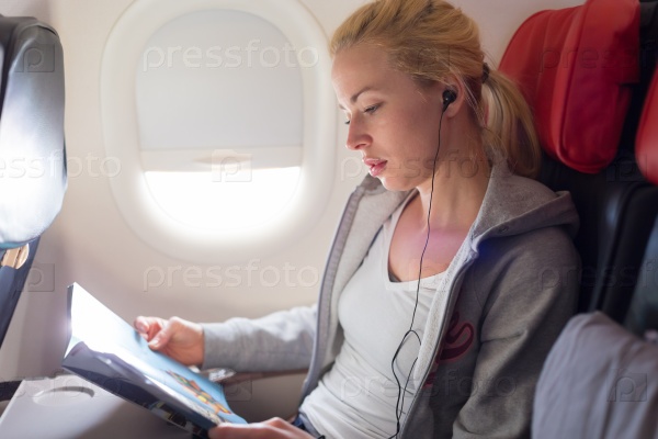 Woman reading magazine and listening to music on airplane. Female traveler reading seated in passanger cabin. Sun shining trough airplane window.