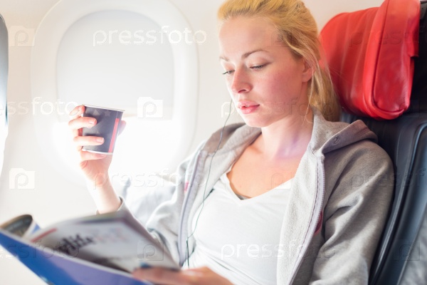 Woman reading magazine and drinking coffeeon airplane. Female traveler reading seated in passanger cabin. Sun shining trough airplane window, stock photo