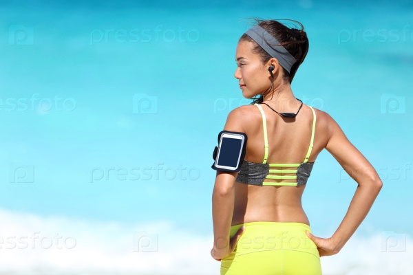 Fitness runner woman with fit back wearing phone armband and wireless headphones on ocean background. Multiracial exercise girl jogging on summer beach with sports smartphone touchscreen for app.