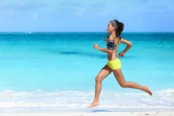 Fitness runner woman running on beach listening to music motivation with phone case sport armband strap. Sporty athlete training cardio barefoot with determination under summer sun.