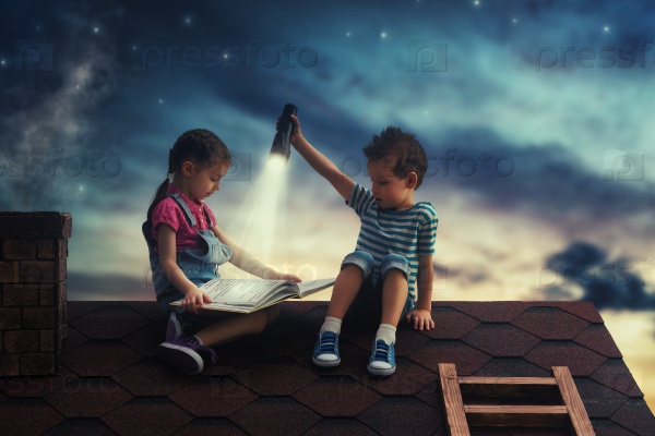 Children reading a book sitting on the roof of the house. Boy and girl reading by the light of a flashlight at night.