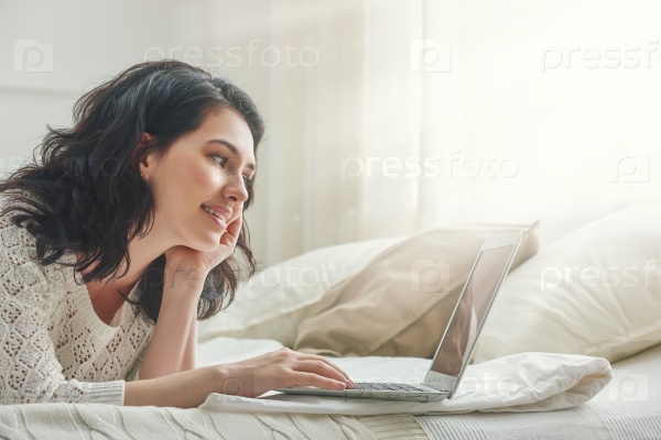 Happy casual beautiful woman working on a laptop sitting on the bed in the house, stock photo