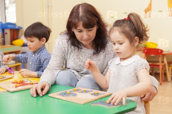 adult beautiful caucasian female teacher collect puzzle with a girl at the green table in the classroom. Horizontal color image