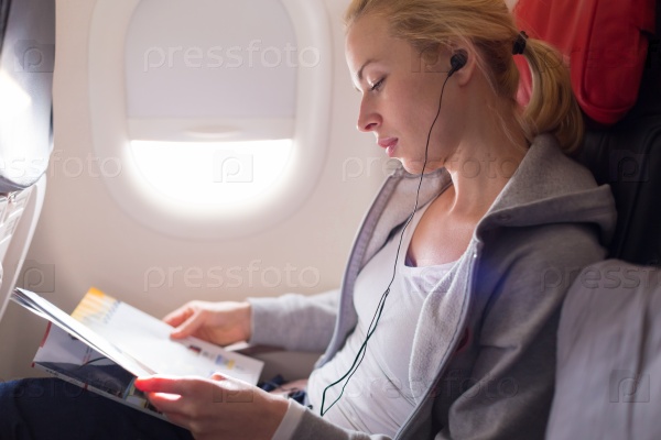 Woman reading magazine and listening to music on airplane. Female traveler reading seated in passanger cabin. Sun shining trough airplane window.