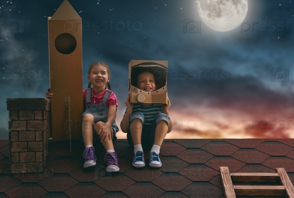 Two little children playing astronauts. Child boy in an astronaut costume and child girl with toy rocket standing on the roof of the house and looking at the sky and dreaming of becoming a spacemen.
