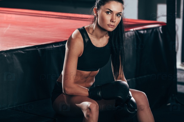 Brunette woman wearing black sportwear and boxing gloves posing near ring, looking at camera, stock photo