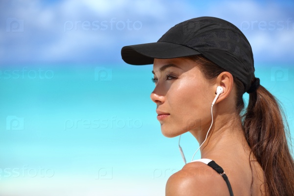 Fitness runner girl listening to smartphone music phone app on beach wearing earphones earbuds and running cap for sun protection. Asian woman healthy and active on summer vacation.
