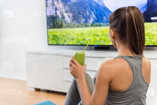 Home woman drinking green smoothie watching tv show on television with weight loss vegetarian diet healthy juice during online fitness dvd workout videos in living room.