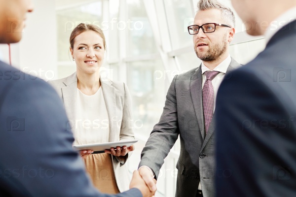 Happy colleagues looking at businessmen handshaking after striking deal, stock photo