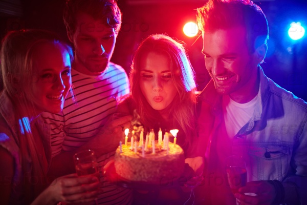 Young woman blowing candles on birthday cake during party with friends, stock photo