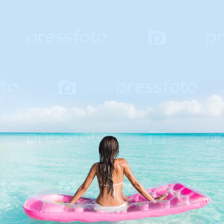 Beach vacation relaxation on ocean water bed. Bikini woman from the back sitting on a pink pool floating air mattress looking at horizon of the perfect turquoise pristine sea in tropical destination.