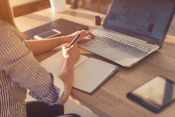 Female designer using laptop, sketching at blank notepad. Woman hand writing in notebook on wooden desk.