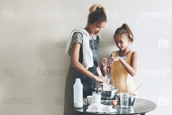 Mom with her 10 years old daughter dressed in linen aprons are cooking together over light wall, lifestyle photo series, stock photo