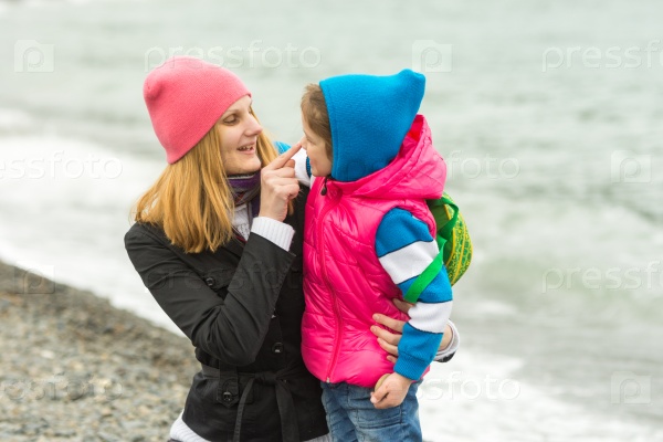 Mother hugging little daughter and fun finger touches her nose at the seaside in cold weather, stock photo