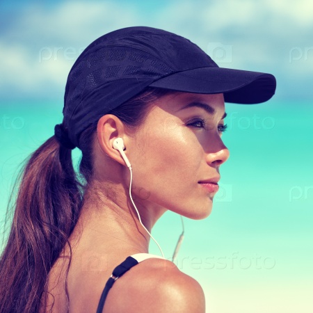 Fitness runner woman listening to music on beach. Portrait of beautiful girl wearing earphones earbuds and running cap for sun protection. Asian woman healthy and active in summer.