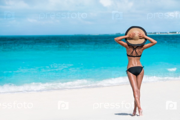 Summer vacation happiness carefree joyful woman standing on white sand enjoying tropical beach destination. Holiday bikini girl relaxing from behind holding straw hat on Caribbean vacation sea water.
