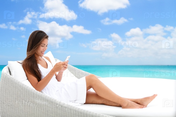 Woman using phone app relaxing on sun bed on outdoor terrace. Home living outside patio furniture Asian girl on smartphone lying on beach lounger in white dress with ocean background enjoying summer.
