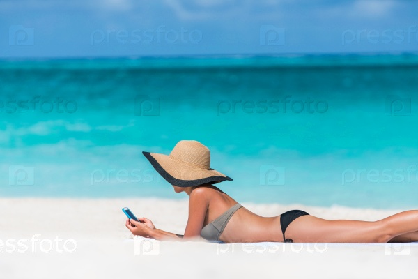 Beach vacation girl using mobile phone app texting sms or browsing on online social media during summer travel holiday. Woman relaxing sunbathing on sand lying on towel wearing sun hat.