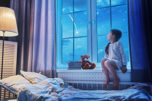 Cute child girl sitting at the window and looking at the stars. Girl making a wish by seeing a shooting star at bedtime night, stock photo