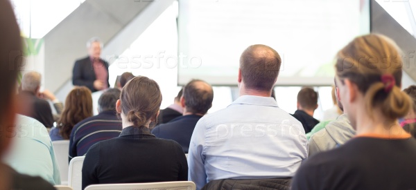 Man giving presentation in lecture hall. Male speeker having talk at public event. Participants listening to lecture. Rear view, focus on people in audience, stock photo