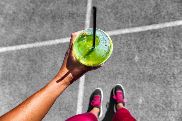 Green smoothie woman drinking plastic cup breakfast meal takeaway to go after morning run on city streets. Healthy lifestyle sporty person pov of hand holding glass with running shoes feet selfie.