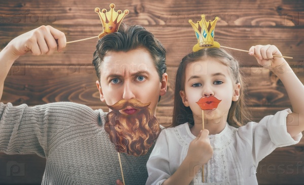 Funny family! Father and his child daughter girl with a paper accessories. Beauty funny girl holding paper lips and crown on stick. Beautiful young man holding paper beard and crown on stick, stock photo