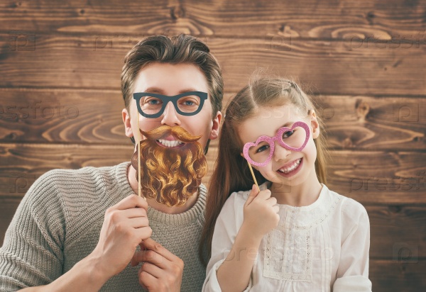 Funny family! Father and his child daughter girl with a paper accessories. Beauty funny girl holding paper glasses on stick. Beautiful young man holding paper beard and glasses on stick.