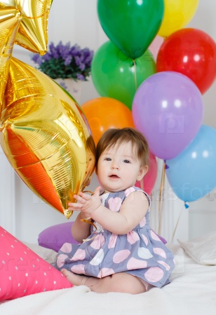Portrait of a little redheaded girl with dark grey eyes, a ridiculous smile, a pink bow on its head, in a beautiful, lush pink dress on a background of Golden stars and colorful balloons, posing in her first birthday on white background